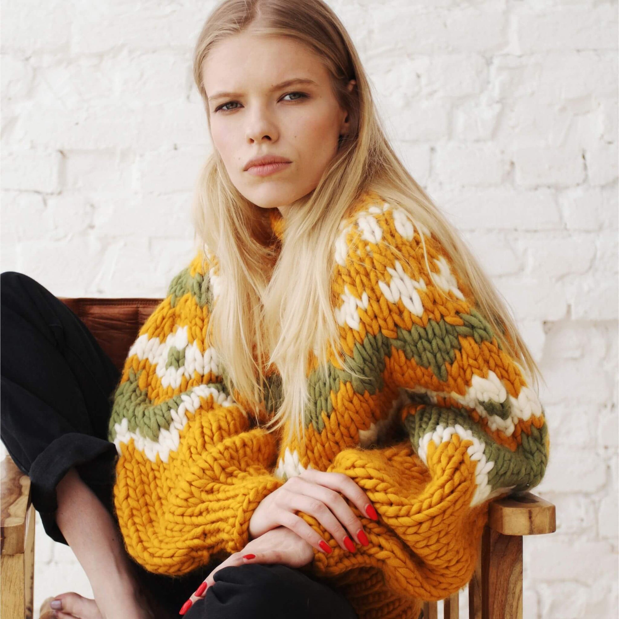 Woman wearing a colorful knitted sweater sitting on a wooden chair against a white brick wall.