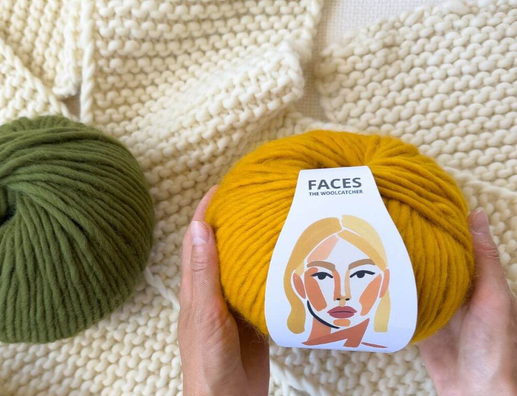 Yellow yarn ball with a 'FACES' label held by hands on a textured beige background